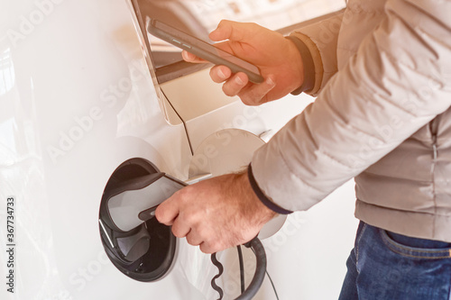 Man charging an electric car and checking the status of battery on this smartphone at sunlight. Car sharing or rental of electric vehicle. 