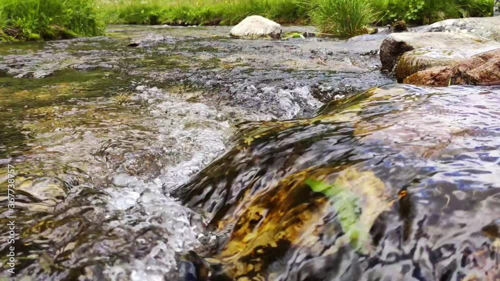 Close-up view mountain stream with stones and sky reflections. Flowing river with smooth stones. Spring water running through stones and green grass Close-up view mountain stream with stones and sky reflections. Flowing river with smooth stones. Spring water running through stones and green grass