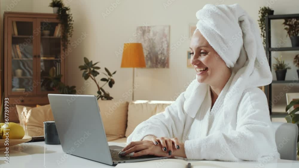 Young cheerful woman in bathrobe and towel on head smiling, waving and talking on video call on laptop while sitting at table in the living room Young cheerful woman in bathrobe and towel on head smiling, waving and talking on video call on laptop while sitting at table in the living room