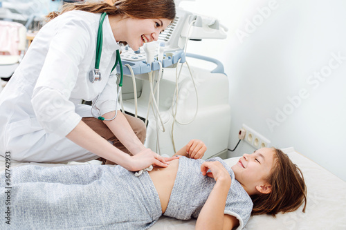 Young female doctor pressing her fingers against stomach of a little girl. She's checking density of her liver. They are smiling at each other.