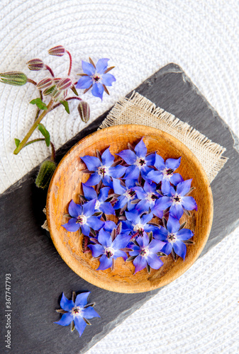 Using beautiful dark blue edible flower for food decoration Borago officinalis (Borago or borage). Flat lay view of blossoms in wood bowl on black stone plate, white background. photo
