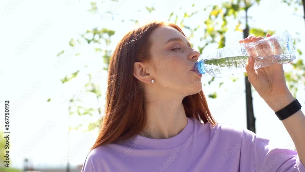 Close-up of beautiful red-haired girl drinking water from bottle outdoors on sunny day. Thirsty young woman with closed eyes drink water outside. Fitness lady enjoying fresh water at cardio training.