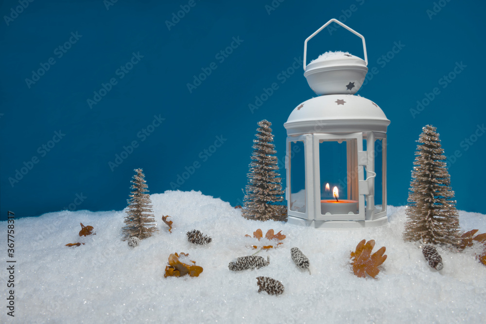 White Christmas lantern on snow with pine cones, small cones, leaves, golden christmas trees and a fir branch in front of dark blue background, copy space