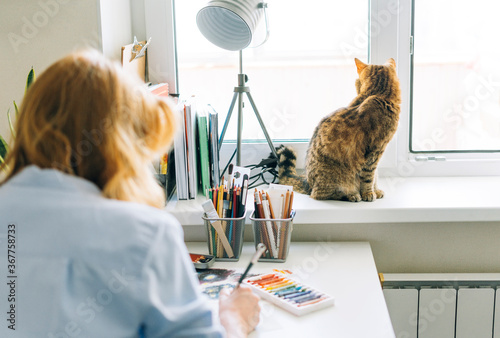 Young woman with red hair illustrator artist draws at desk at home. Cat sitting on the window sill