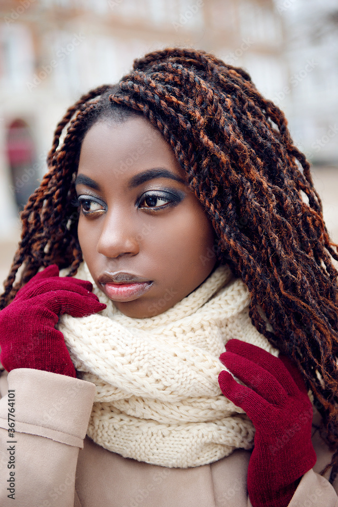 African girl in a coat and with make-up holds hands in gloves near a scarf on a winter city street, close-up. African girl in a coat and with make-up holds hands in gloves near a scarf on a winter city street, close-up.