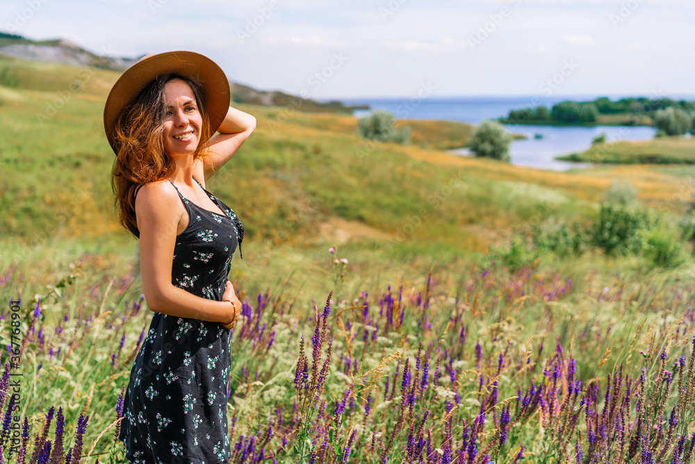 A happy young woman in a dress and hat stands against a background of blooming wildflowers and tall grass in a hilly area