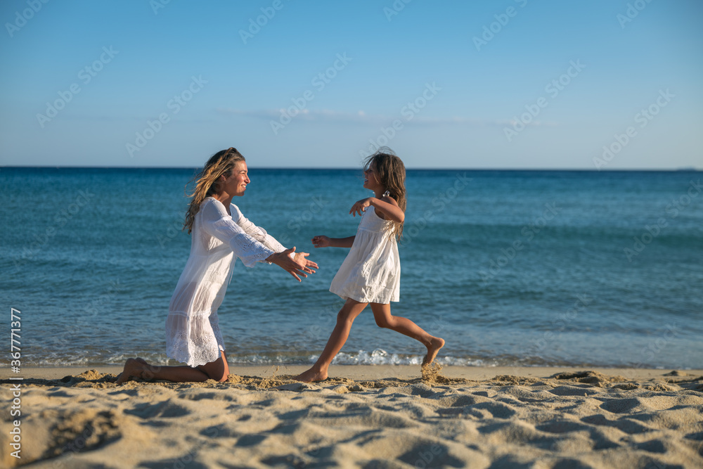 Authentic shot of happy little daughter is running to give a hug and kiss with affection to a mother on a seaside beach during holiday vacation at sunset.