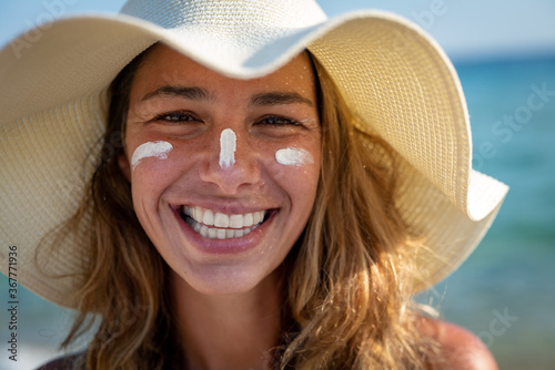 Close up of happy young smiling woman with straw hat and sunscreen or sun tanning lotion on her face to take care and protect skin on a seaside beach during holidays vacation. photo