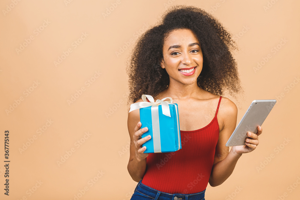 Happy birthday holiday concept. Happy african american lady in casual laughing while holding present gift isolated over beige background. Using tablet. Happy birthday holiday concept. Happy african american lady in casual laughing while holding present gift isolated over beige background. Using tablet.
