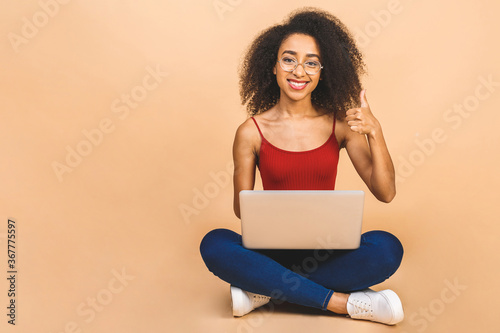 Portrait of smiling young afro american black woman using laptop while sitting on a floor with legs crossed isolated over beige background. Thumbs up.