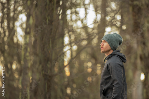 Side view of a man in the forest wearing gray hoddie and beanie with a winter forest as background