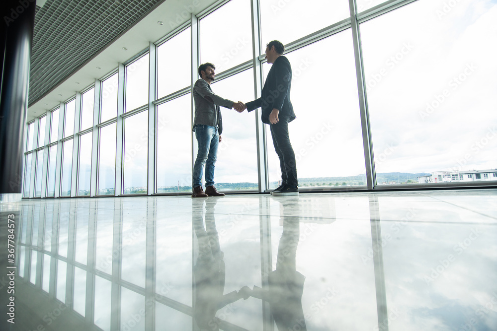 Full length side view of businessmen shaking hands in office building windows background Full length side view of businessmen shaking hands in office building windows background