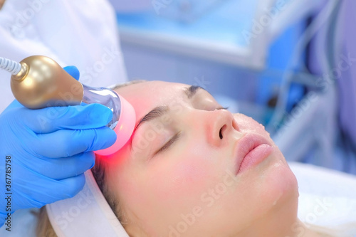 Cosmetologist doctor making phonophoresis procedure on woman face with hyaluronic acid gel, side view. Beautician moving manipula with hot red light on face. Apparatus procedure in cosmetology clinic.