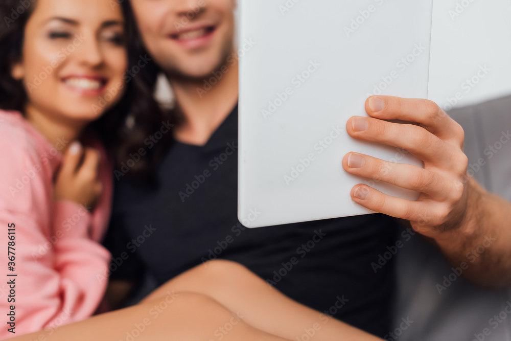 Selective focus of man holding digital tablet near cheerful girlfriend on couch isolated on grey Selective focus of man holding digital tablet near cheerful girlfriend on couch isolated on grey