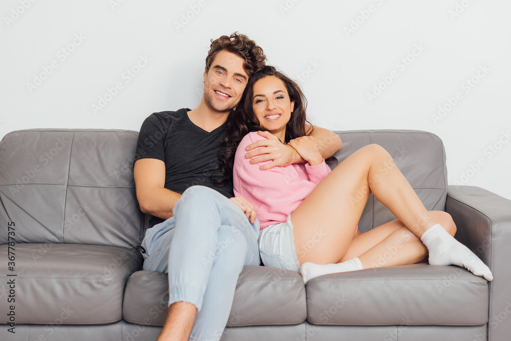 Handsome man hugging cheerful girlfriend on sofa on grey background