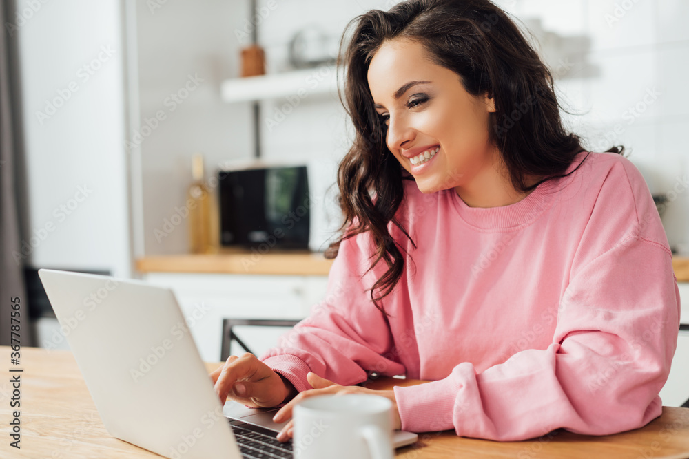 Selective focus of smiling freelancer using laptop near cup of coffee on kitchen table Selective focus of smiling freelancer using laptop near cup of coffee on kitchen table