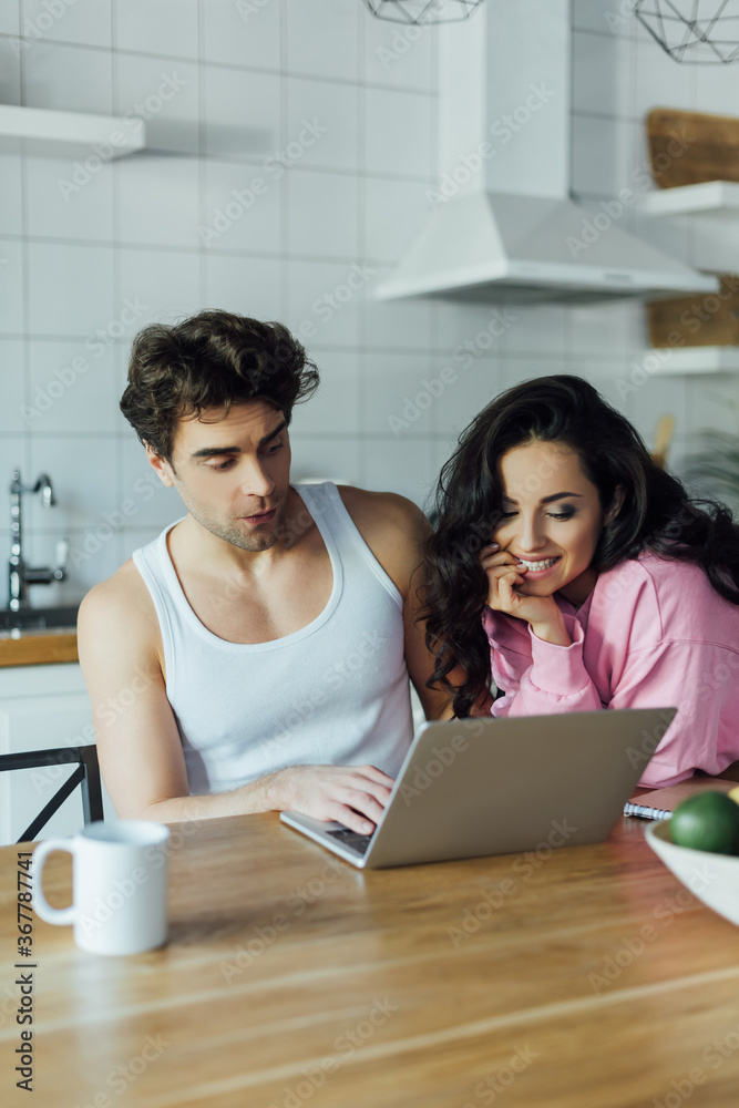 Selective focus of smiling woman looking at laptop near boyfriend in kitchen