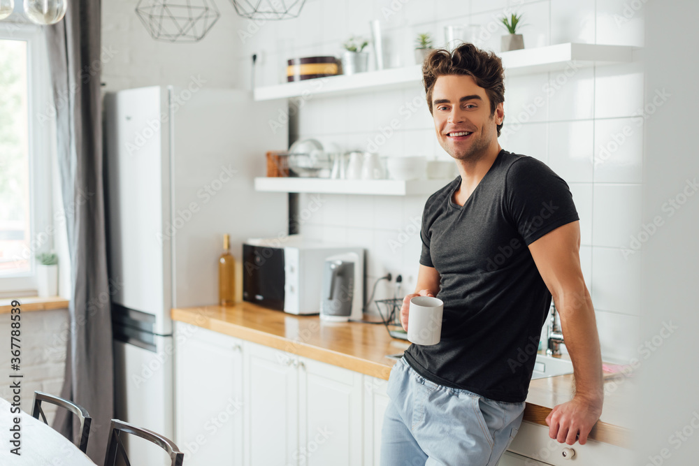 Selective focus of handsome man smiling at camera while holding cup of coffee near worktop in kitchen
