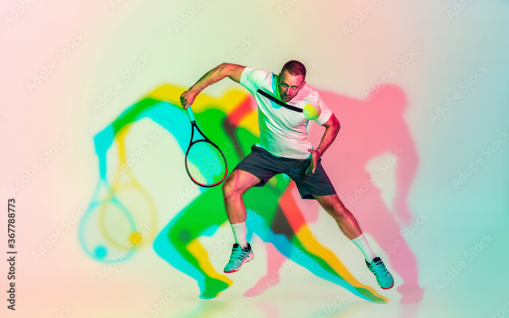 In jump. One caucasian man playing tennis on studio background in colorful neon light. Fit young professional male player in motion or action in sport game. Concept of movement, sport, healthy