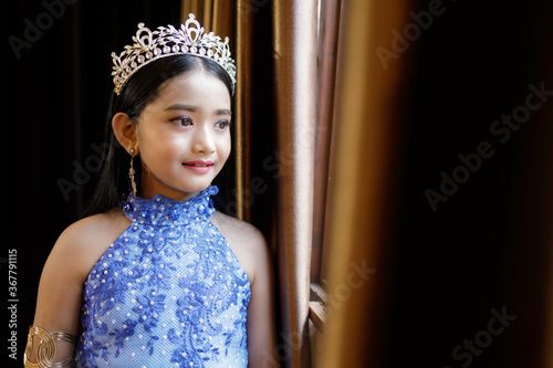 A portrait of a cute Asian girl, makeup, wearing a blue evening dress and a crown standing by the window.