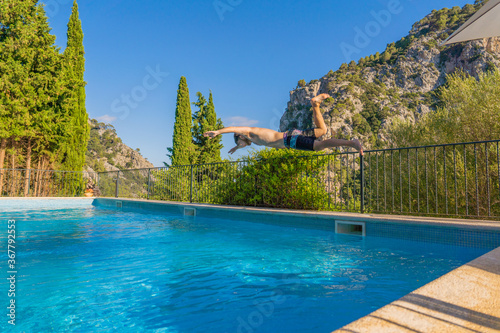 fair-skinned blond boy jumping into the pool on his head on a terrace with mountains and a Mediterranean landscape with pine trees in the background  mid-day sun and clear ground.