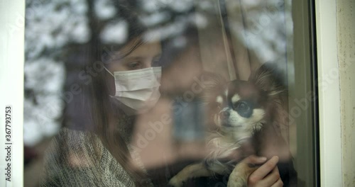 Teen age girl wearing protecting mask looks through the window handeling a dog, wishing of playing outside. Sad child plays with her pet. Quarantine makes children sad,pets suffer from extra attention photo