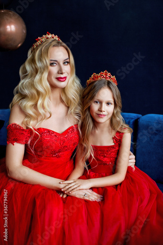 Portrait of mother and daughter in luxurious red dresses and diadems. Mom touches her daughter's hair © popovatetiana
