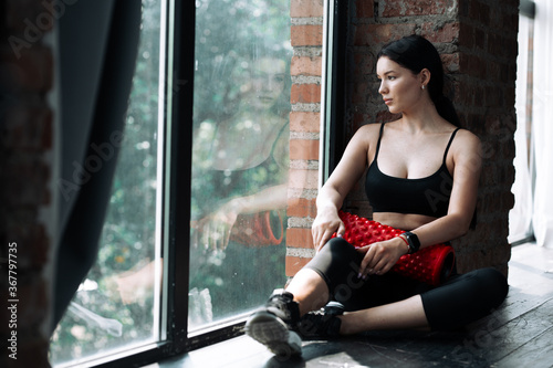 An Asian woman with black hair and a black tank top sits near a panoramic window and rests after a workout with a red massage cylinder to work out the fascia of the muscles of the body