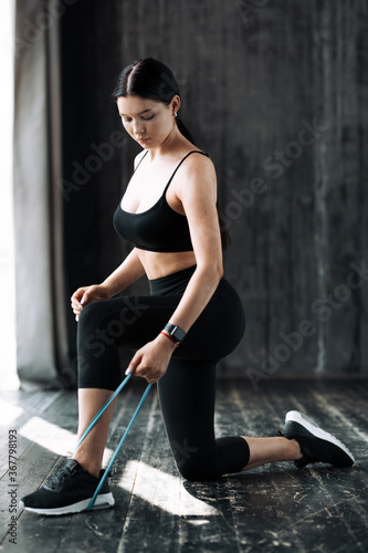 Full-length A young woman is kneeling and pressing down an elastic expander with her leg to pump up the muscles of her arms on the floor in a loft