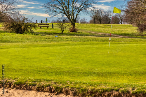 Putting Green mit gelber Fahne im Vordergrund ein Sandbunker im Hintergrund zwei Golfspieler photo