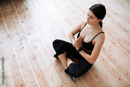 Top view of meditating yoga coach in black sportswear holding hands in front of him and sitting on wooden floor in lotus position