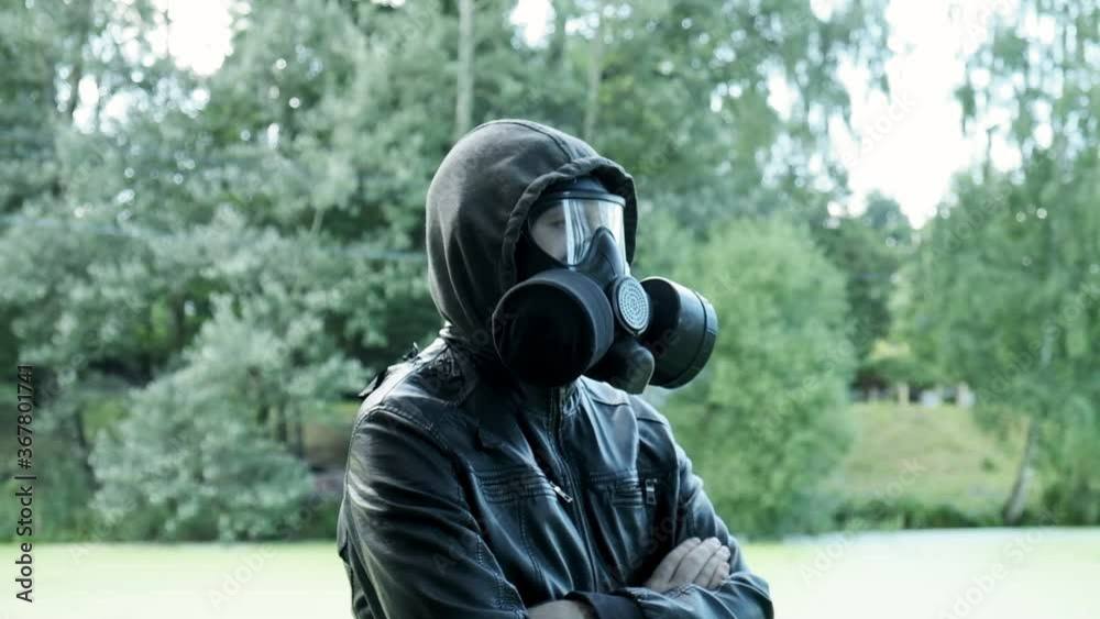 man in gas mask, black leather jacket with hood standing outdoors at forest near lake. protection from chemical weapons, virus epidemic. Post apocalyptic world. survivor, stalker after nuclear war man in gas mask, black leather jacket with hood standing outdoors at forest near lake. protection from chemical weapons, virus epidemic. Post apocalyptic world. survivor, stalker after nuclear war