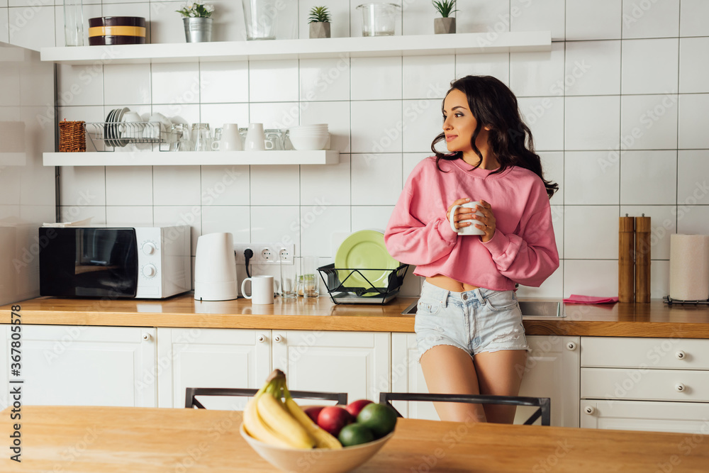 Selective focus of beautiful brunette woman holding cup of coffee in kitchen