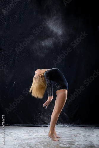 Beautiful plump blonde girl wearing a black gymnastic bodysuit covered with clouds of the flying white powder jumps dancing sitting on a dark. Artistic conceptual and advertising photo.