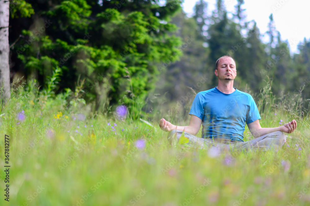 Obraz premium Man is meditating on green grass in the nature with face raised up to sky and eyes closed on sunny summer day. Concept of meditation, dreaming, wellbeing and healthy lifestyle.