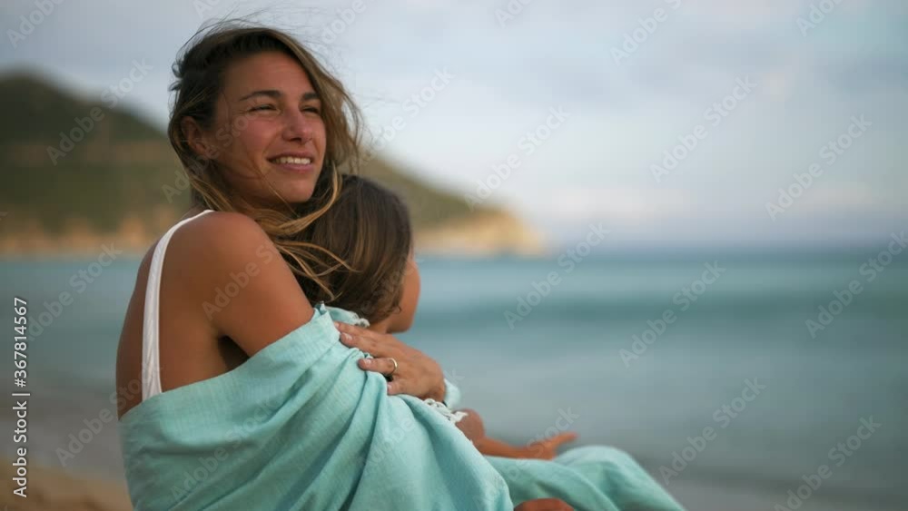 Authentic shot of an young carefree happy smiling mother and little daughter embraced under a striped cover are enjoying their time together on a seaside at sunset during holiday vacation. Authentic shot of an young carefree happy smiling mother and little daughter embraced under a striped cover are enjoying their time together on a seaside at sunset during holiday vacation.