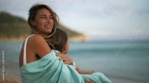 Authentic shot of an young carefree happy smiling mother and little daughter embraced under a striped cover are enjoying their time together on a seaside at sunset during holiday vacation.  photo