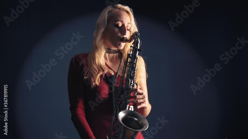 Young beautiful woman in red concert dress playing a melody on saxophone over dark background in studio photo