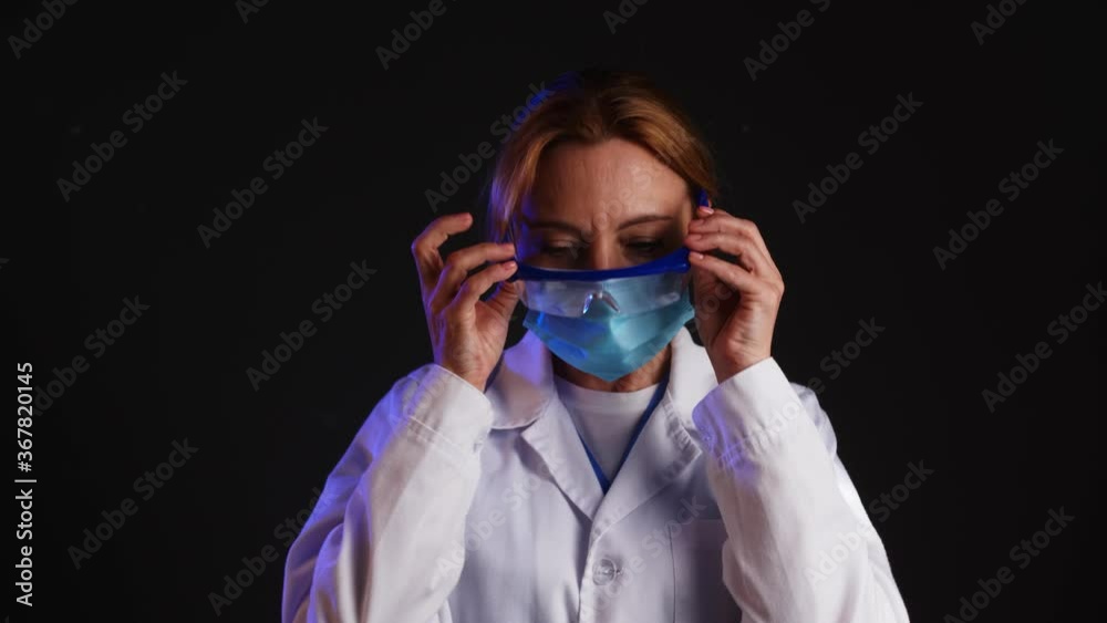 Tired female doctor taking off protective medical hat, eyeglasses and mask against dark background Tired female doctor taking off protective medical hat, eyeglasses and mask against dark background