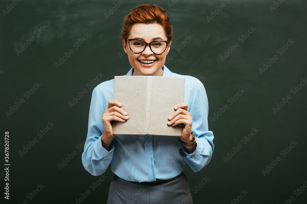 attractive, happy teacher in eyeglasses holding open book while standing near chalkboard attractive, happy teacher in eyeglasses holding open book while standing near chalkboard