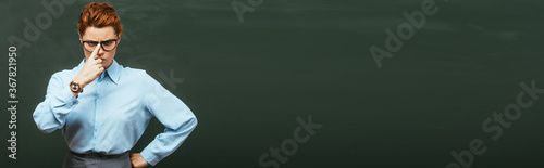 horizontal concept of strict teacher touching eyeglasses while standing with hand on hip near chalkboard photo
