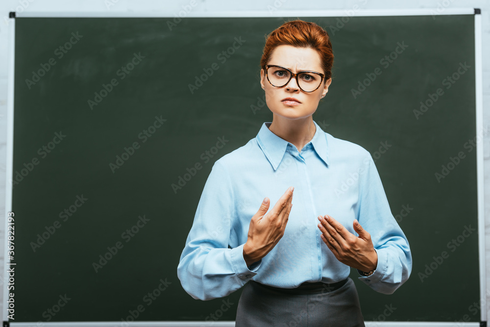 serious teacher in eyeglasses looking at camera while standing near chalkboard
