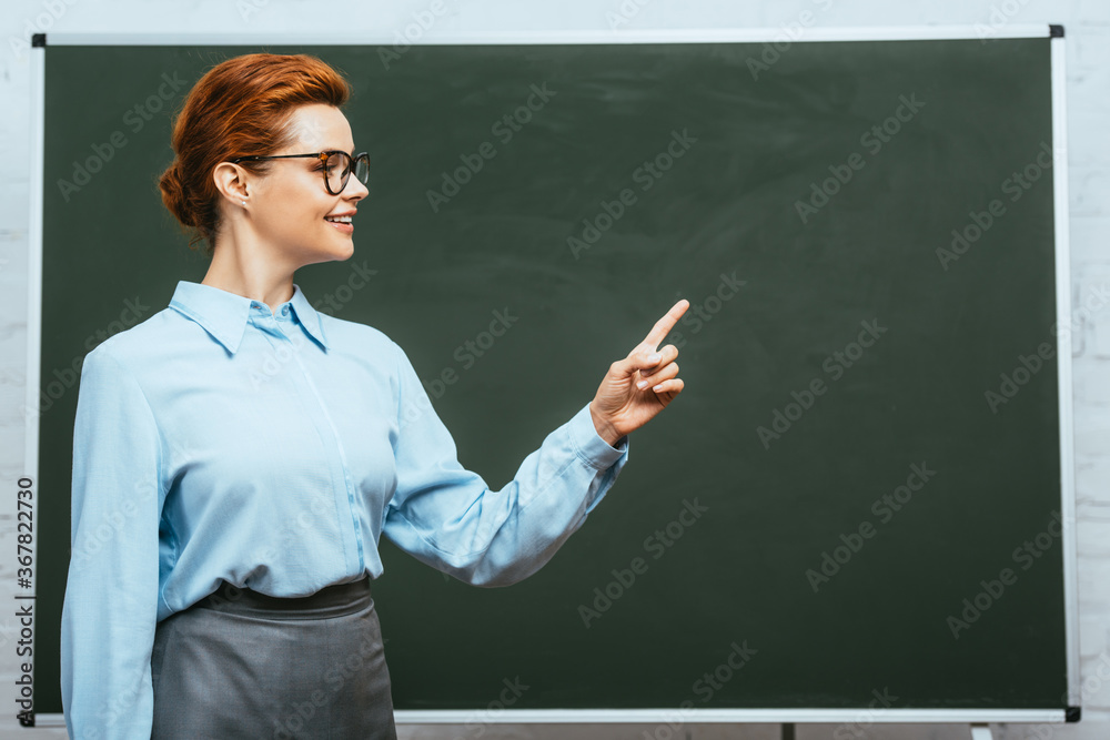 smiling teacher looking away and pointing with finger near chalkboard