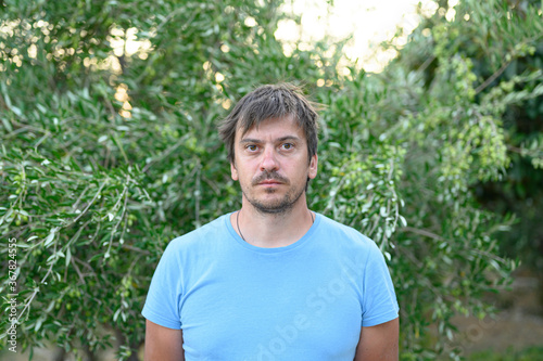 portrait of a serious tired young 30+ year old tourist, unshaven and shaggy, against the green plant olive tree in the nature outdoor