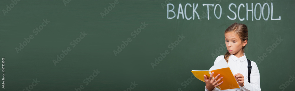horizontal image of surprised schoolgirl looking at copy book near chalkboard with back to school lettering horizontal image of surprised schoolgirl looking at copy book near chalkboard with back to school lettering