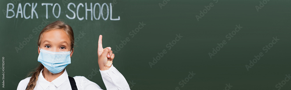 horizontal image of schoolgirl in medical mask pointing with finger near chalkboard with back to school inscription - obrazy, fototapety, plakaty 
