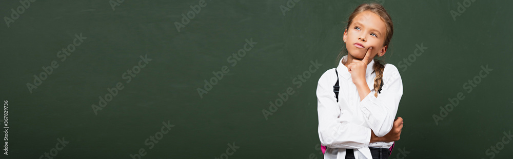 horizontal concept of thoughtful schoolgirl touching face and looking up near chalkboard