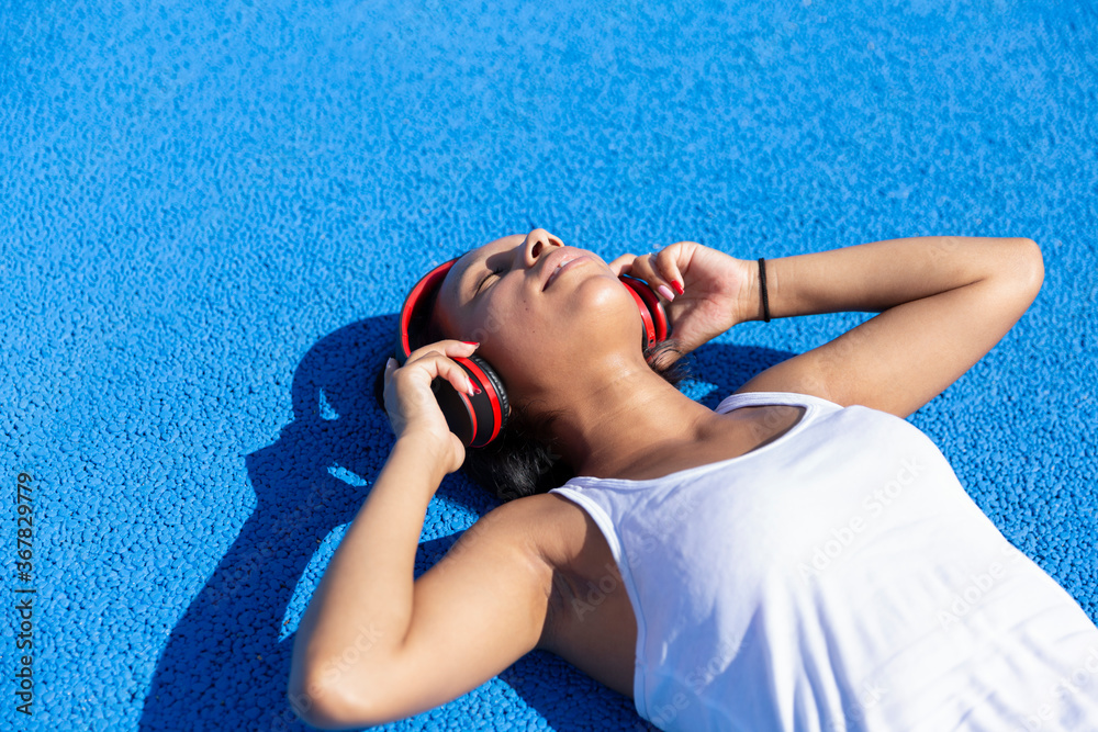 Portrait of a young brunette girl lying on a blue athletic track listening to music through her headphones. Space for text. Concept of healthy life.