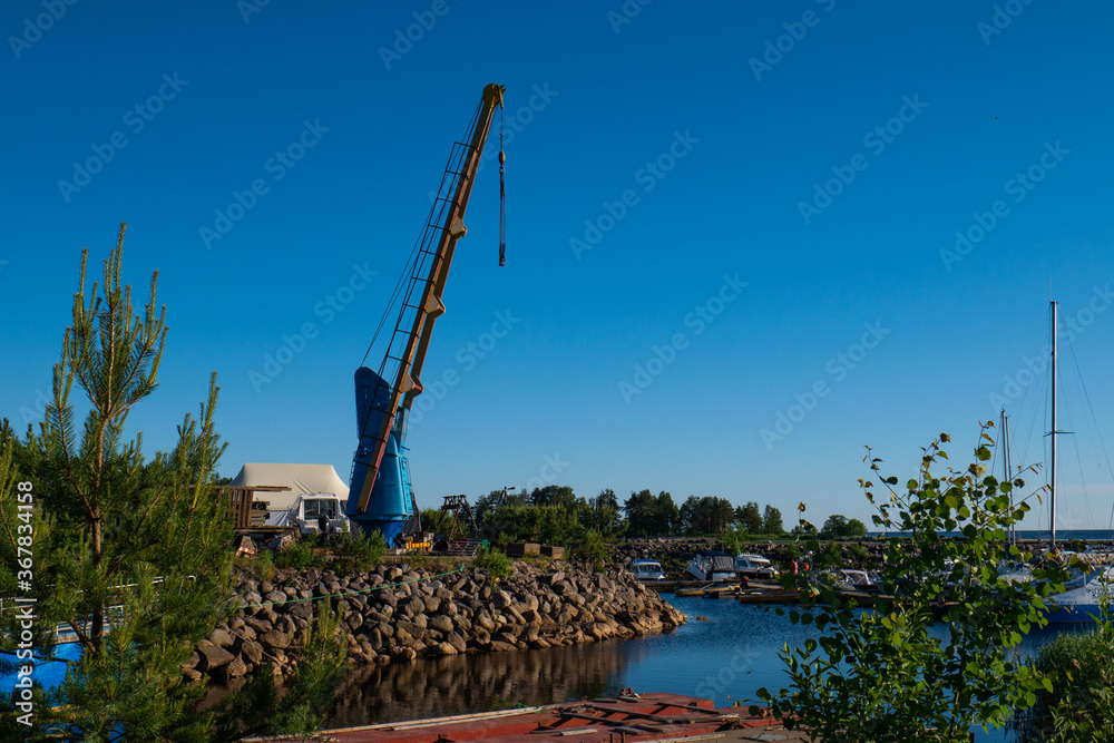 A high crane for yachts against the blue sky and stone shore. Blue-yellow crane for boats.