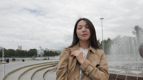 A girl with an Asian appearance in a brown trench stands against the fountain in the center of the city and enjoys a summer evening. High quality 4k footage